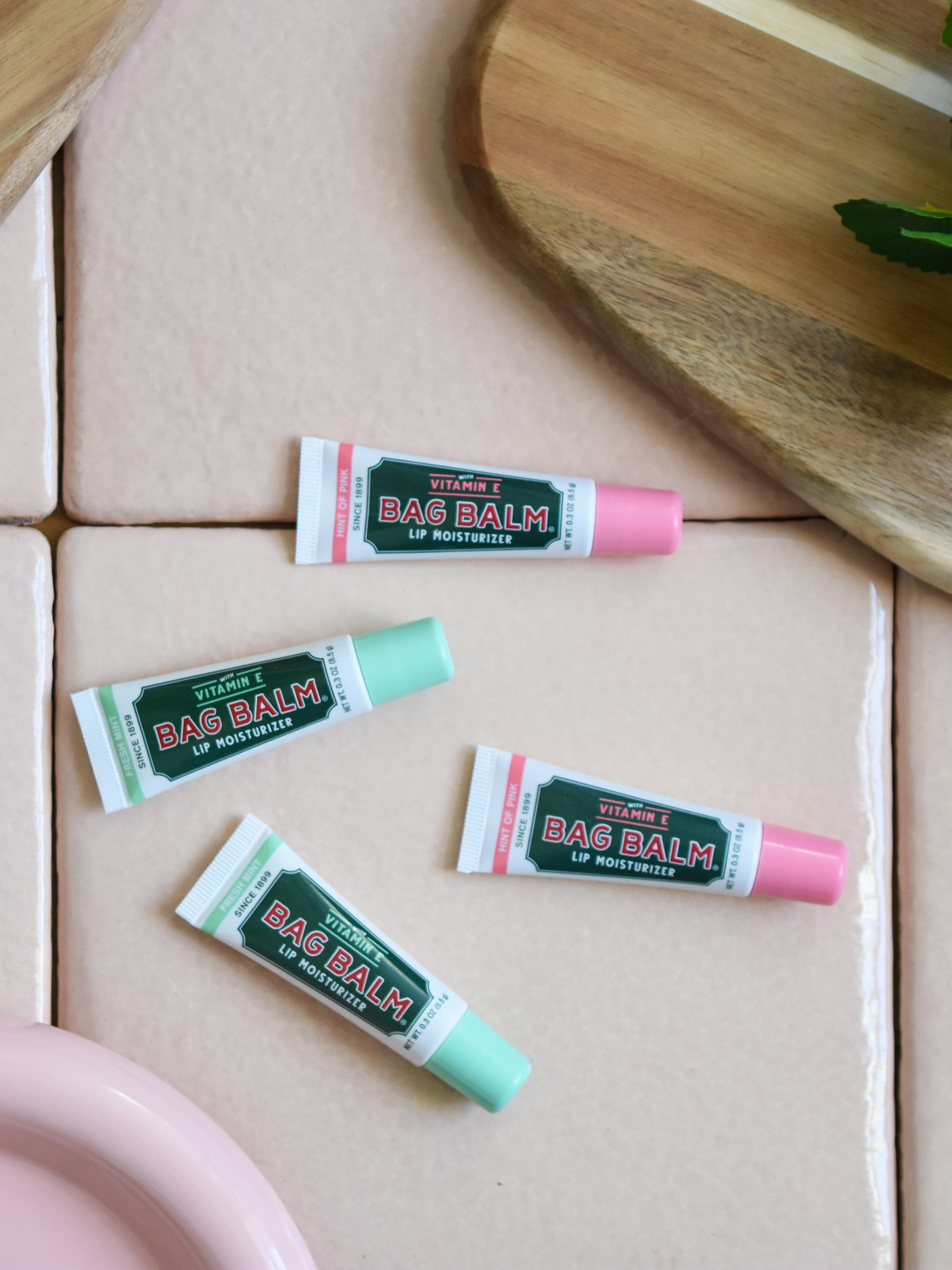 Hint of Pink Lip Balm and Fresh Mint Lip Balm flat lay on pink tile with wood and ceramic plates