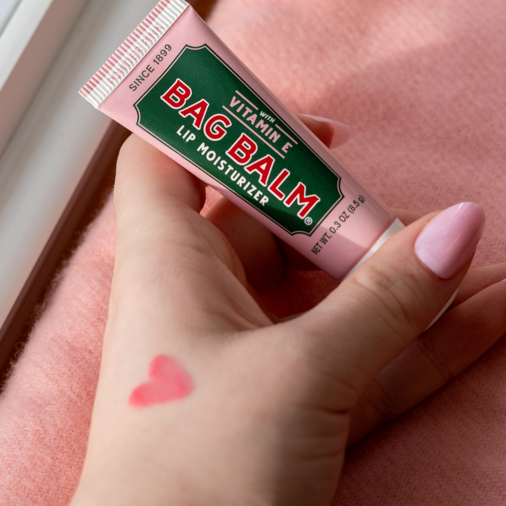 Hand holding a tube of Bag Balm Powder Pink lip balm with a heart-shaped swatch on skin.