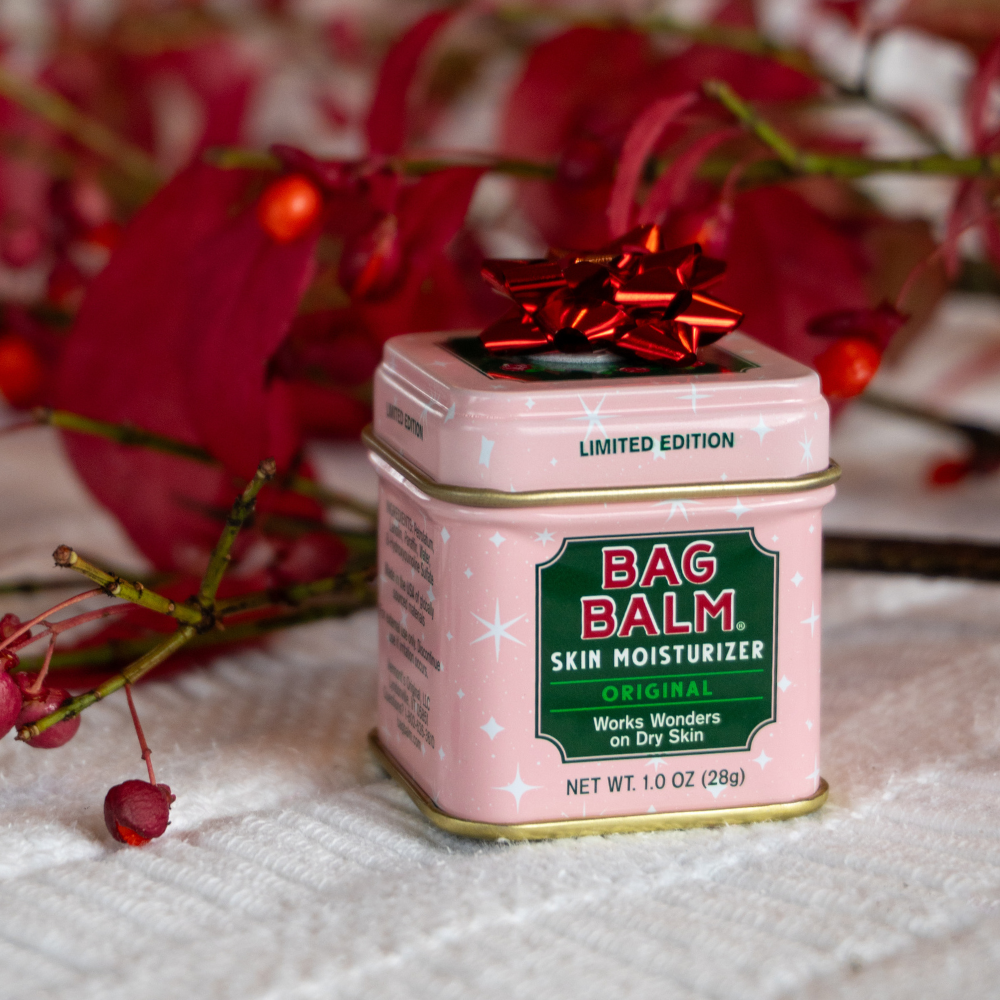 Pink tin of Bag Balm skin moisturizer with a red bow, surrounded by red flowers on a white surface.