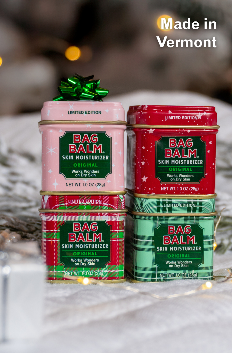 Four tins of Bag Balm skin moisturizer in red, green, and pink on a white surface with blurred lights in the background.