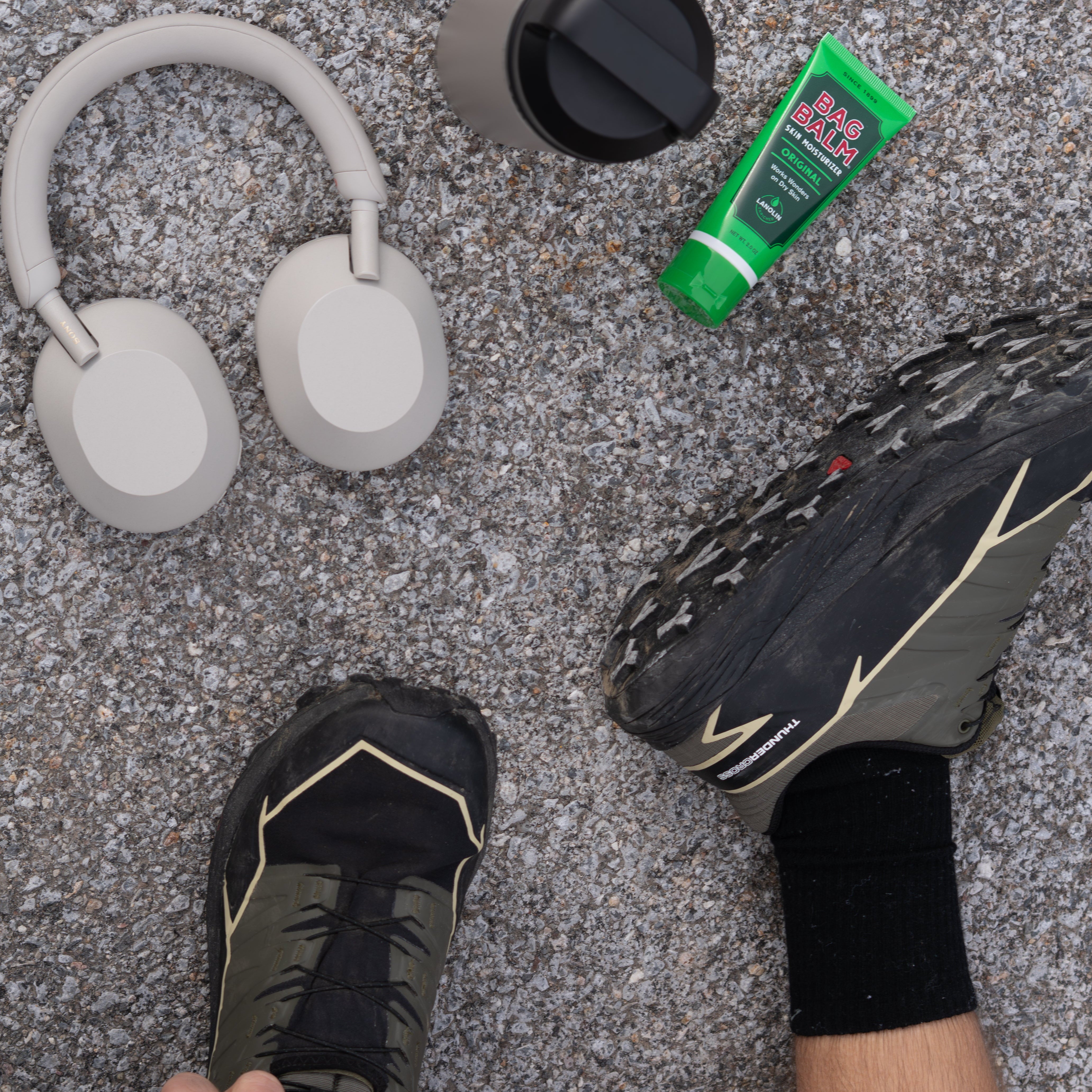 Person wearing running shoes with a bottle of Bag Balm and headphones on pavement