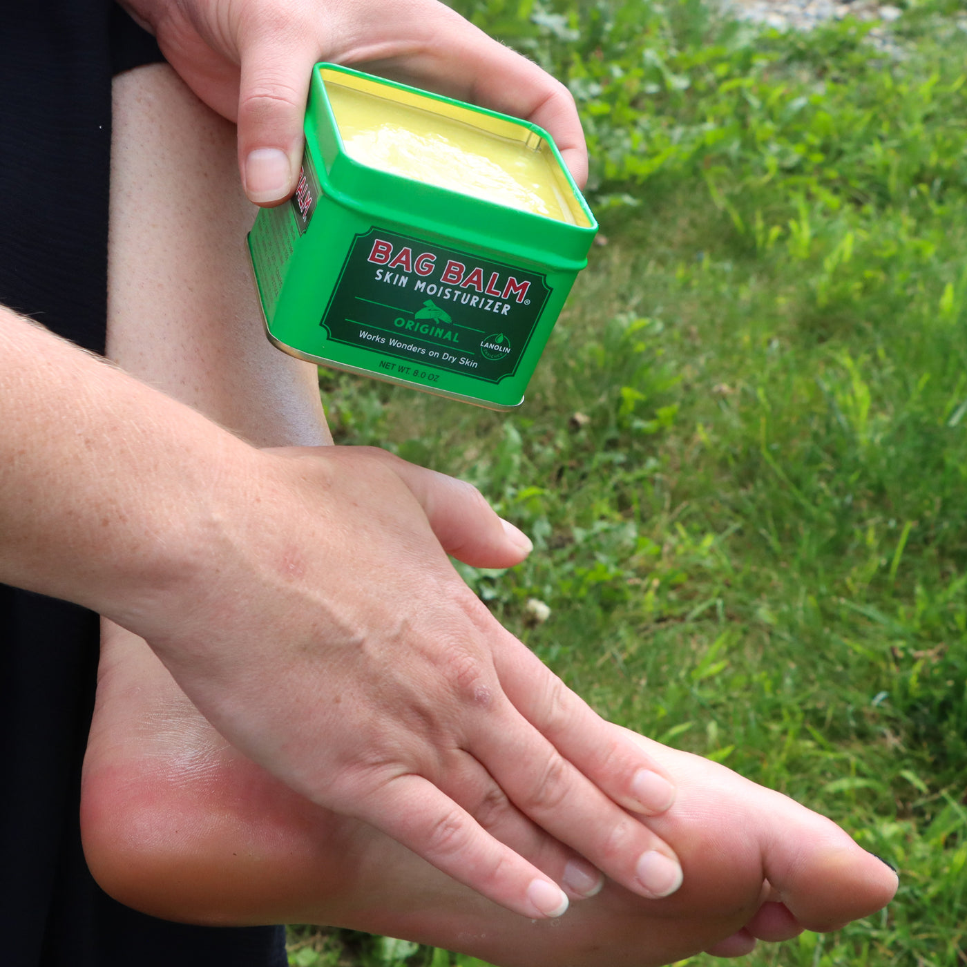 A person holding a green tin of Bag Balm and applying it to their feet
