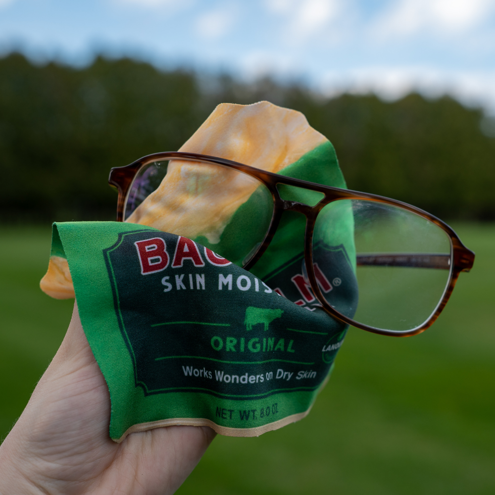 A glasses wipe shaped like a Bag Balm tin wiping a pair of glasses outside