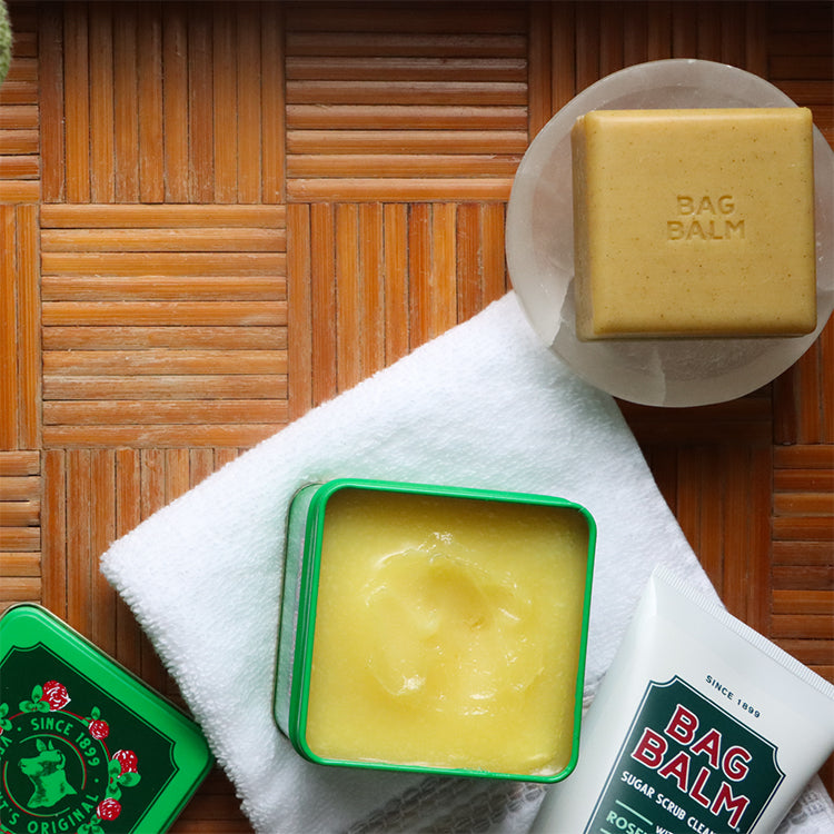 Bag Balm 8 oz tin, open on a white washcloth displaying the ointment inside. The Bag Balm Sugar scrub is in the bottom right, just visible in the shot. A light brown bar of soap with the bag balm logo is in the top right.