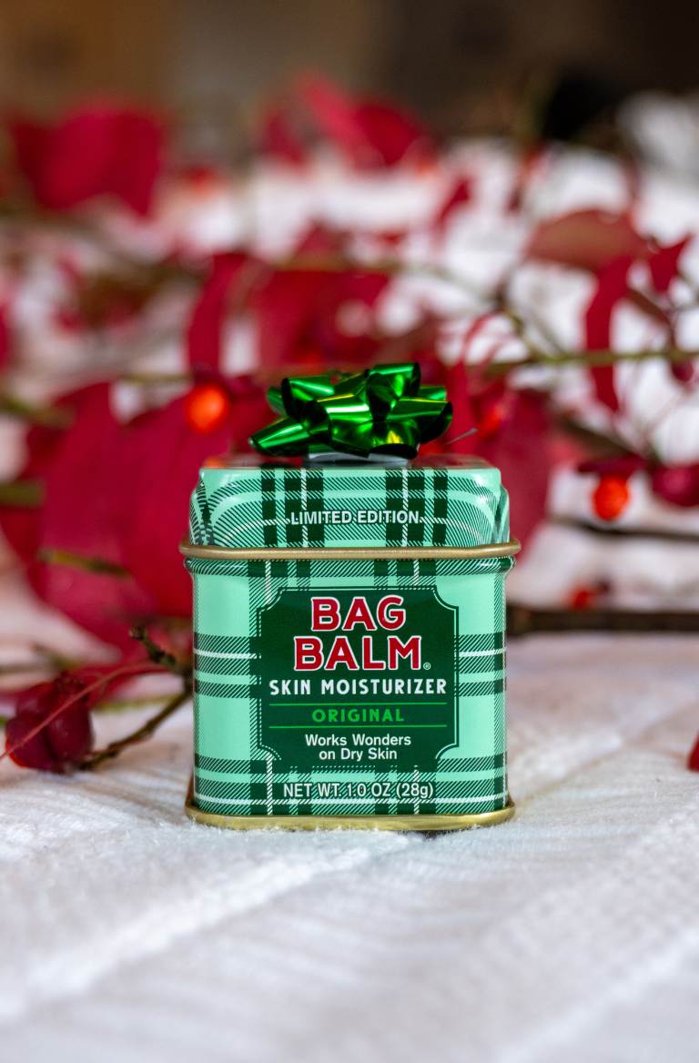 Green tin of Bag Balm skin moisturizer with a green bow on a white surface with red foliage in the background.