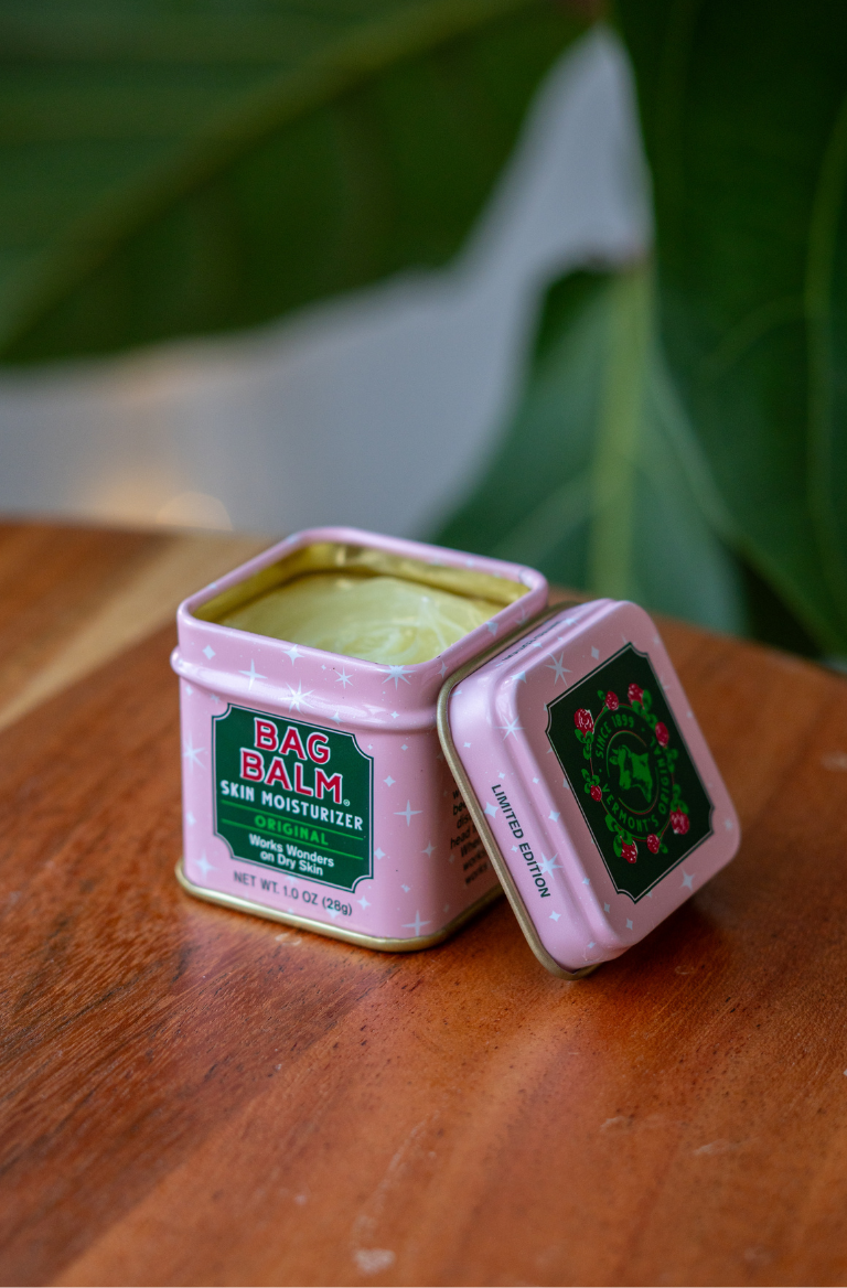 Pink tin of BAG BALM skin moisturizer on a wooden surface with green leaves in the background