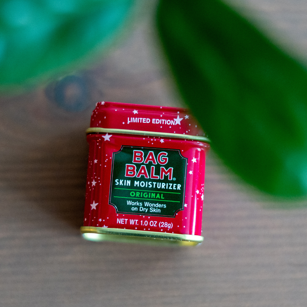 Red tin of Bag Balm skin moisturizer on a textured surface with green leaves in the background