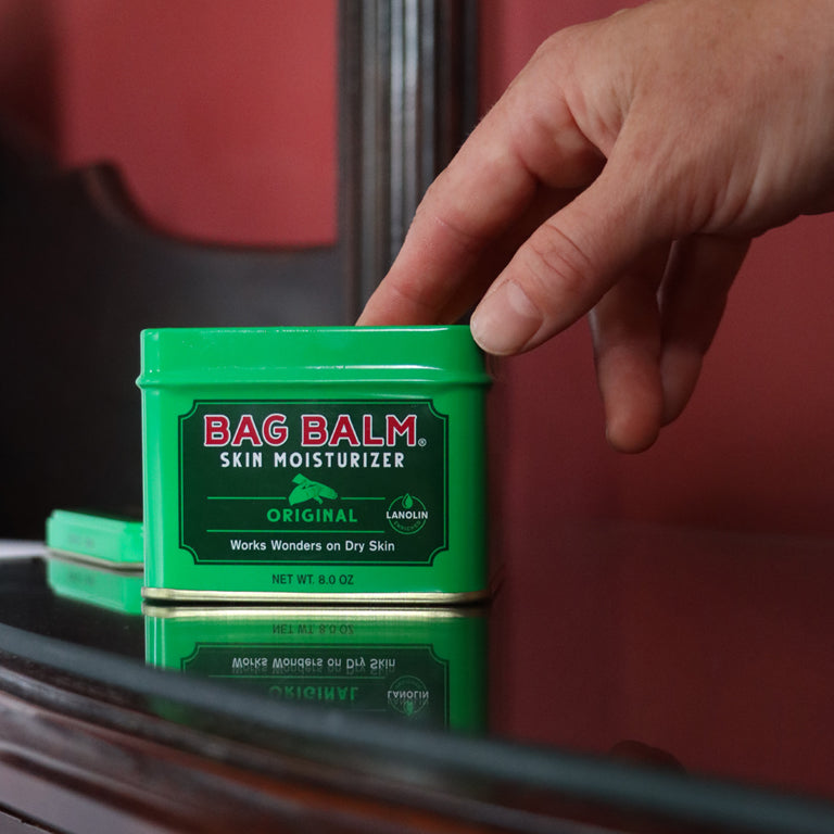 An open tin of green Bag Balm and a person's hand reaching into the container