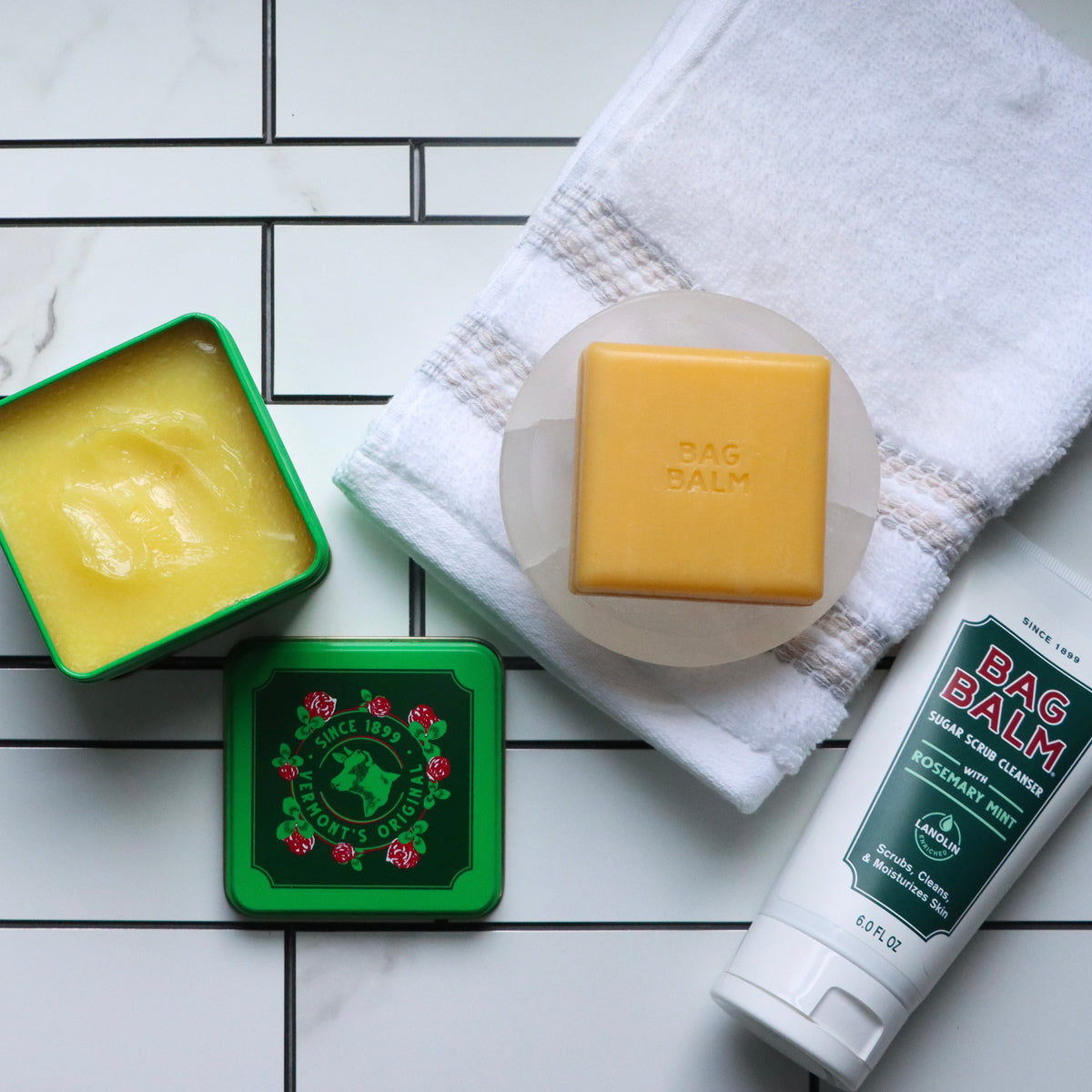 A green tin of open Bag Balm, a bar of Bag Balm soap, and Bag Balm Sugar Scrub against a white tile floor