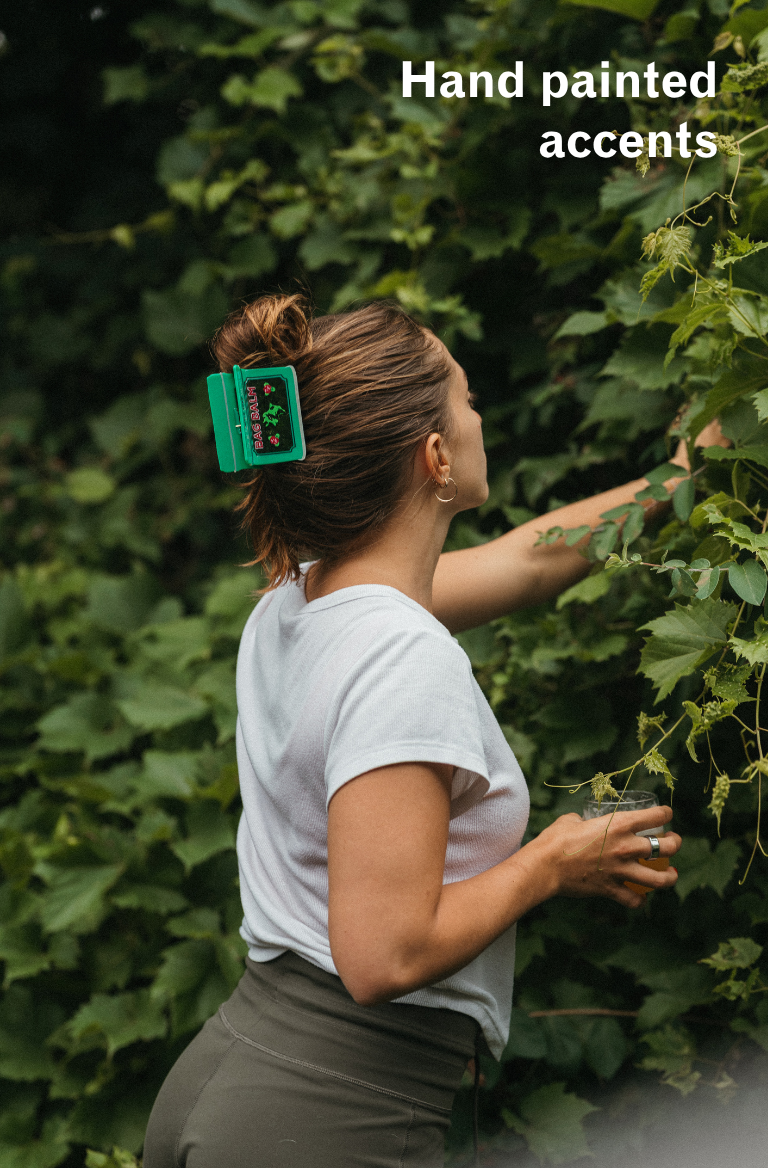 A woman in a white t-shirt and olive green pants stands outdoors among lush green ivy, holding a small glass. She has a bright green rectangular Bag Balm tin shaped hair clip holding her hair in a bun.