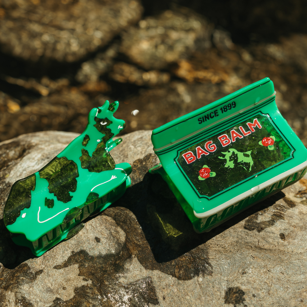 A green cow-shaped Hair claw clip and Bag Balm tin-shaped hair claw clip placed on a rocky outdoor surface with blurred natural background.