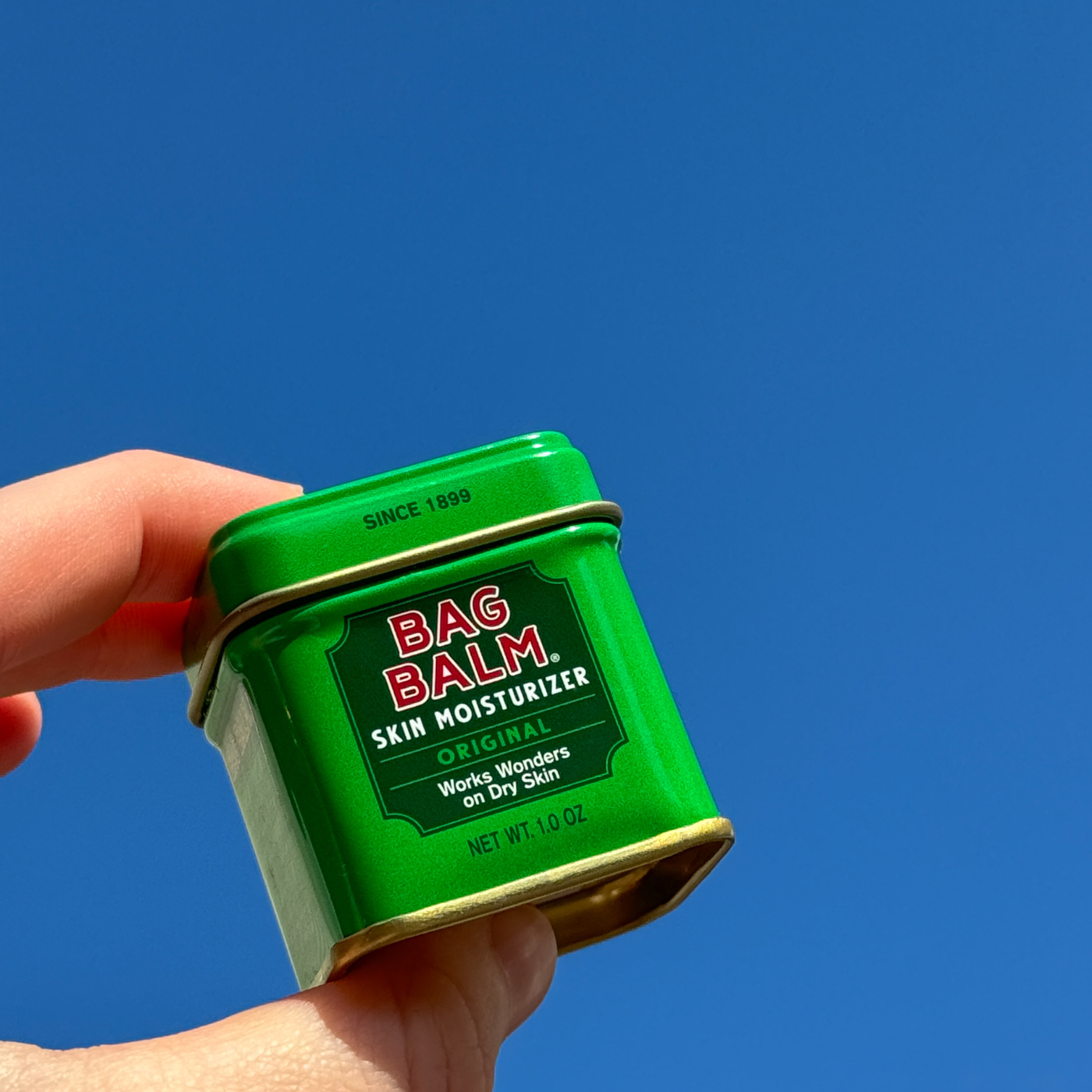 A 1 oz green tin of Bag Balm set against a blue sky