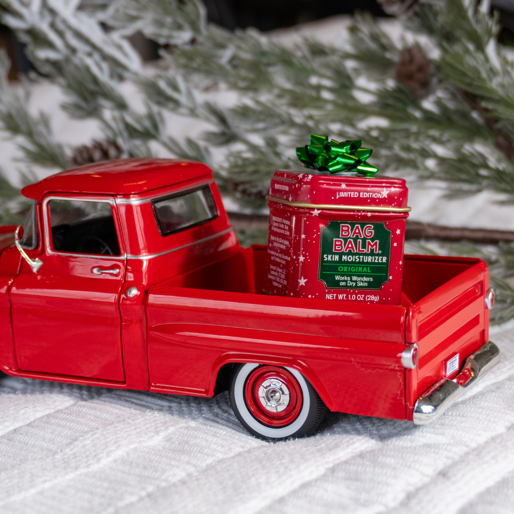 Red toy truck with a mini tin of Bag Balm in the bed, against a Christmas tree background.