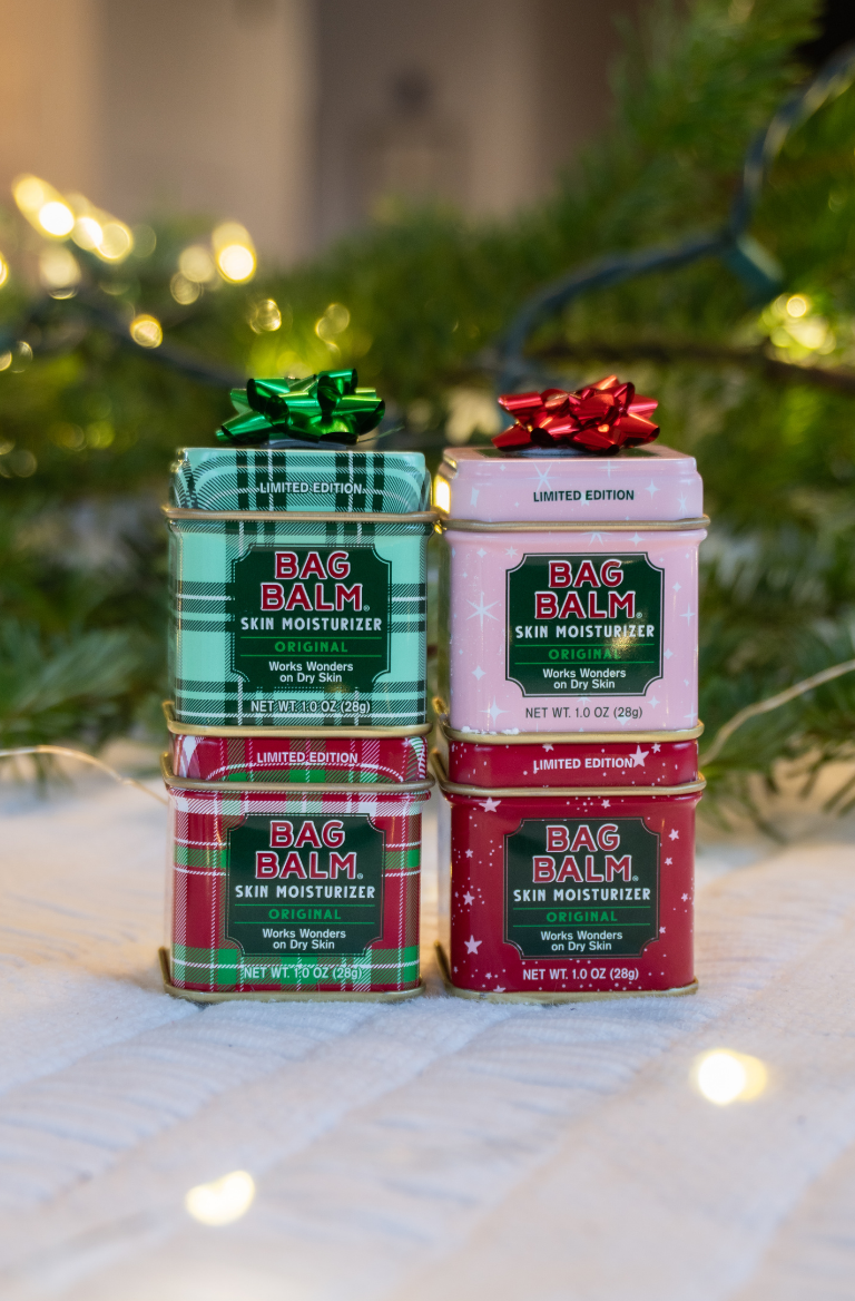 Stack of Bag Balm tins with decorative bows on a white surface with blurred greenery in the background.