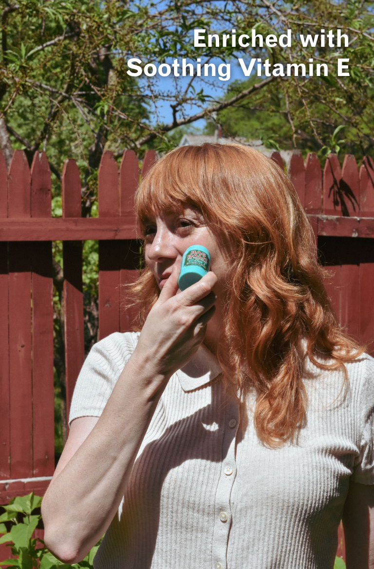 Woman applying Bag Balm Stick outdoors with text 'Enriched with Soothing Vitamin E'.