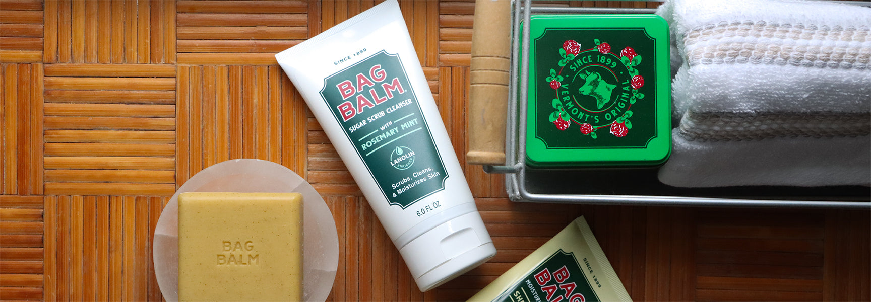 An image containing the following three Bag Balm products on a wood background: Bag Balm tin, Moisturizing Bar Soap, Sugar Scrub Cleanser
