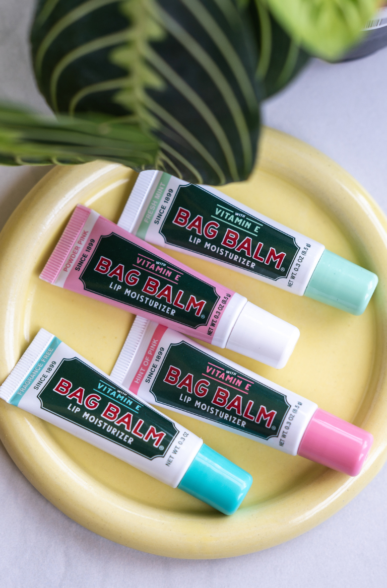 Four tubes of Bag Balm Lip Moisturizers on a yellow plate with a leafy plant nearby.