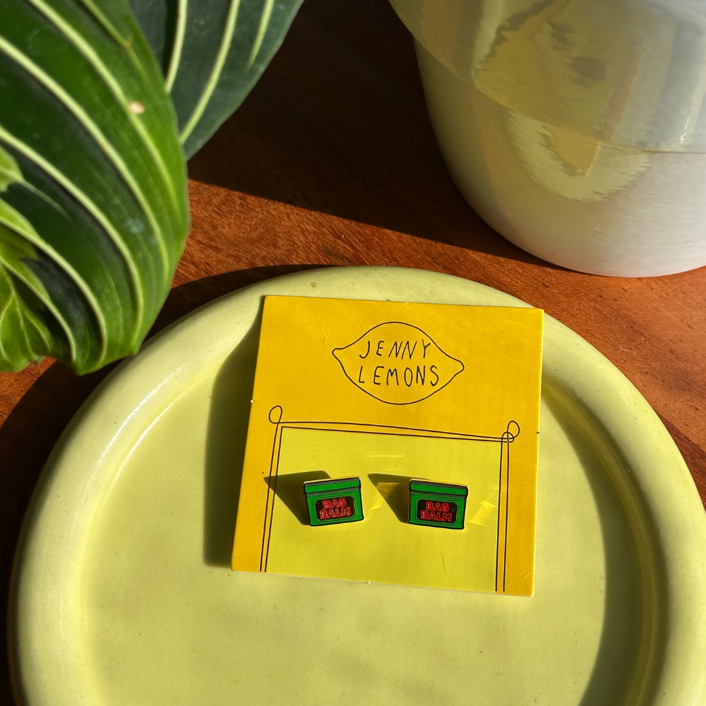 A pair of small rectangular earrings displayed on a yellow backing card that reads 'JENNY LEMONS.' The earrings are green with red details, placed on a pale yellow plate on a wooden surface with a plant leaf and white ceramic object in the background.